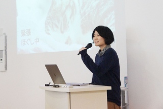 写真：マイクを持って話す小林さん。背景のスライドは猫の写真に「緊張ほぐし中」の文字